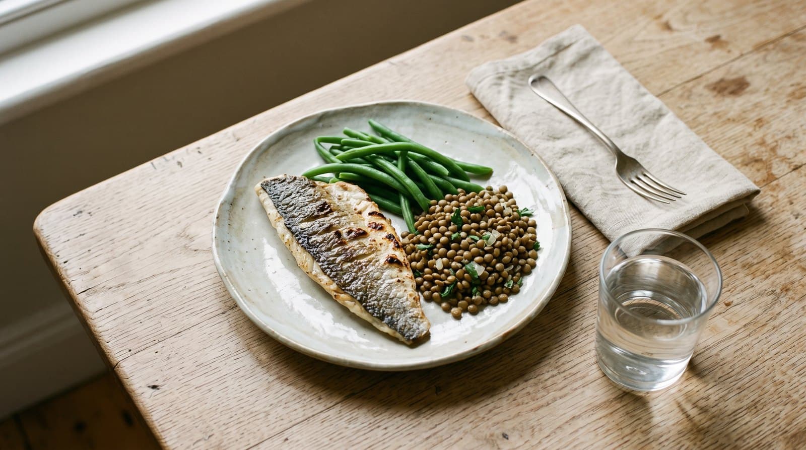 Repas simple sur une table en bois clair : filet de poisson grillé, haricots verts, lentilles et verre d'eau