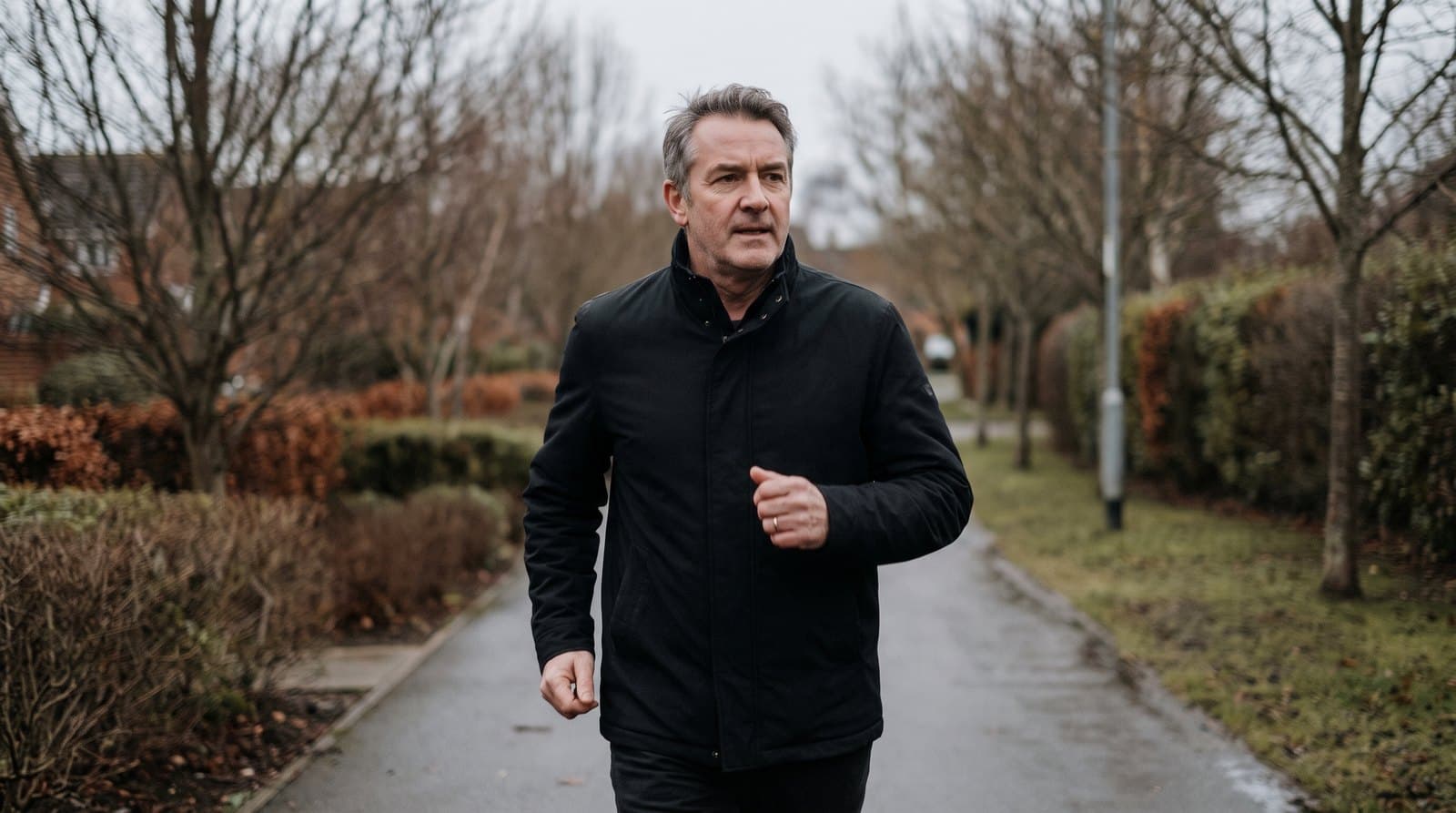 Homme d'une cinquantaine d'années marchant d'un pas soutenu sur un chemin pavé, en veste sombre et chaussures de sport ordinaires